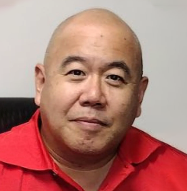 A bald man is wearing a red shirt and sitting in a chair.