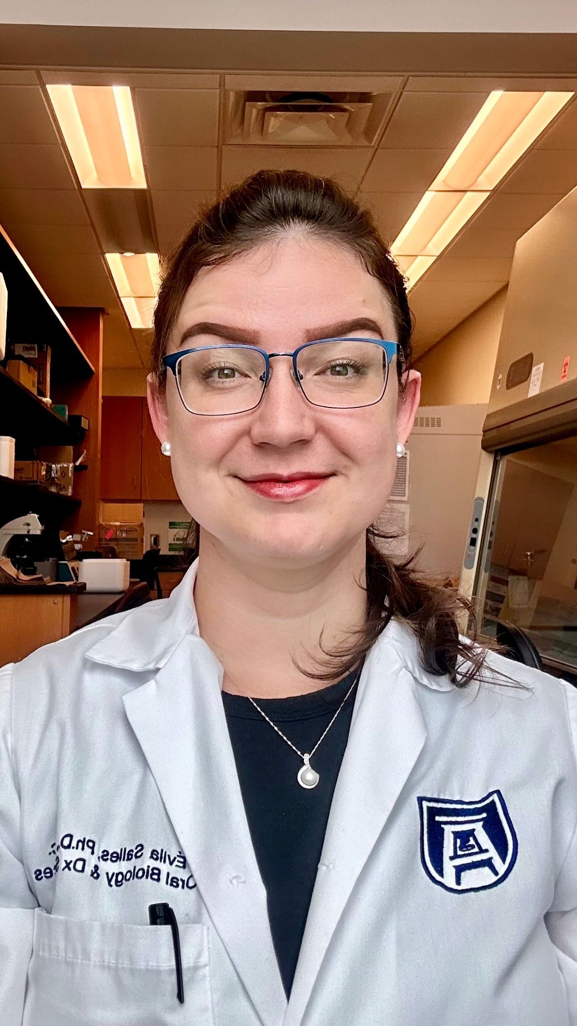 A woman wearing glasses and a lab coat is smiling in a lab.