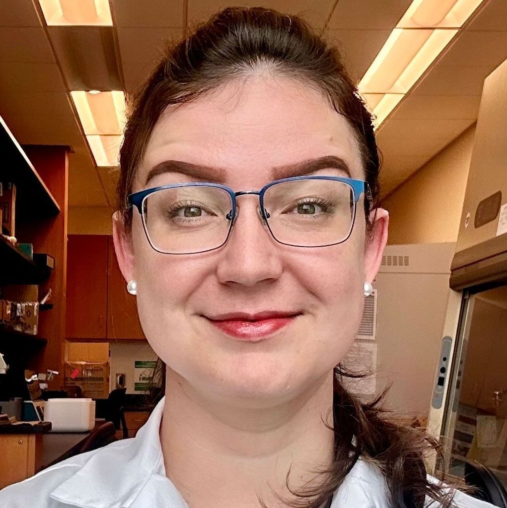 A woman wearing glasses and a white shirt is smiling in a lab.