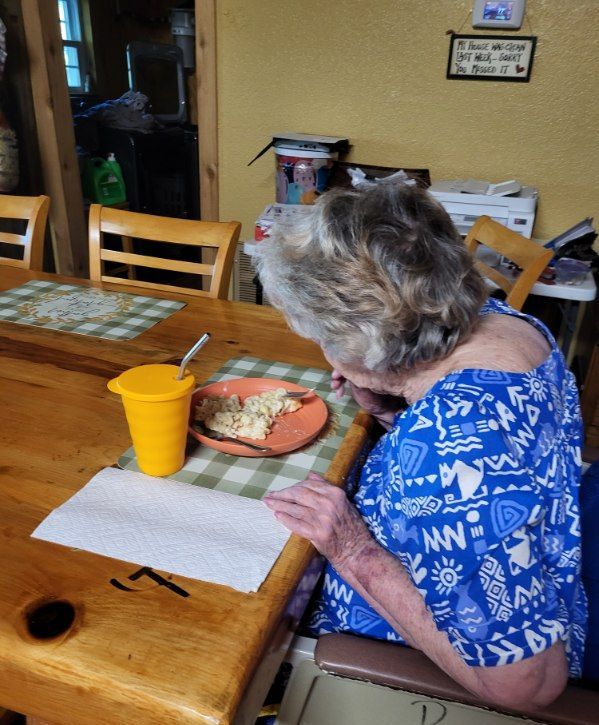 An elderly woman sits at a table with a plate of food and a yellow cup