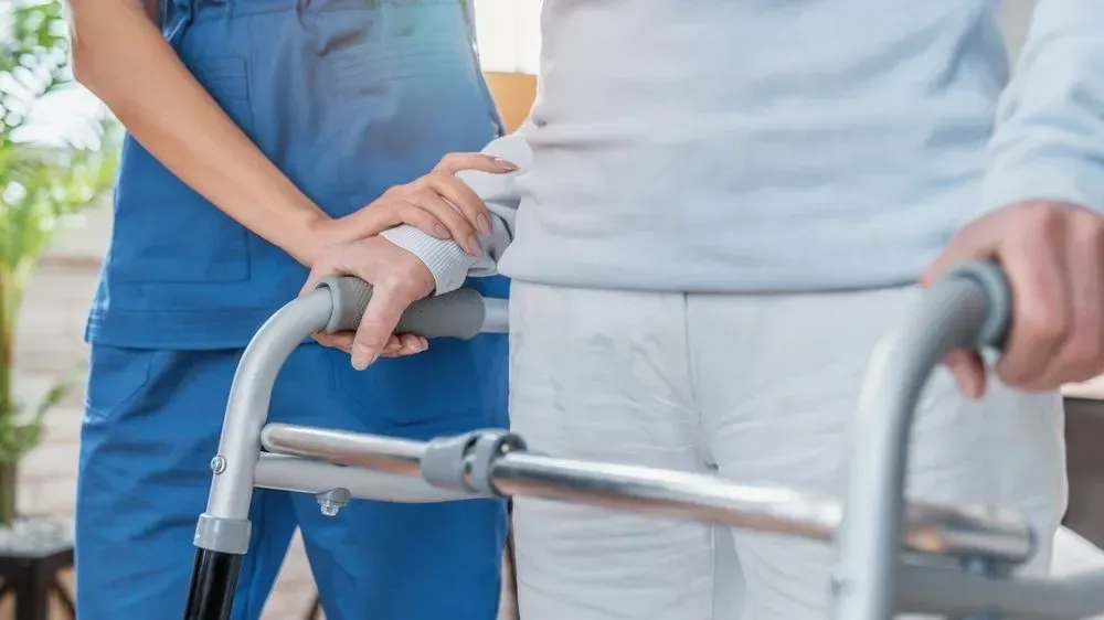 A caregiver in blue uniform assists an elderly person using a walker, indoors.