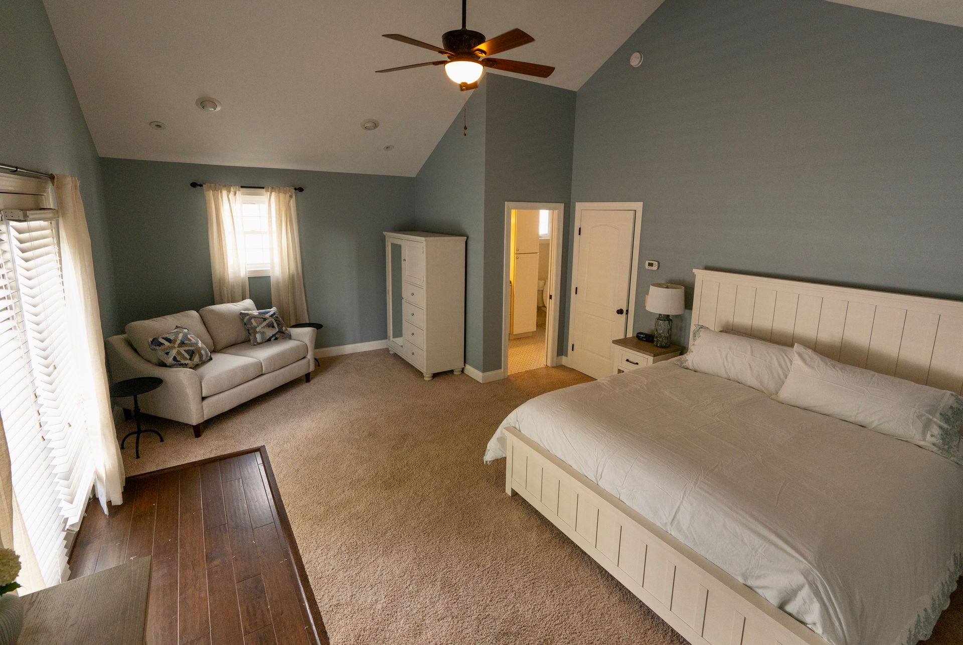 Spacious bedroom with a bed, couch, wardrobe, and bathroom. Gray walls, brown carpet, and a ceiling fan.