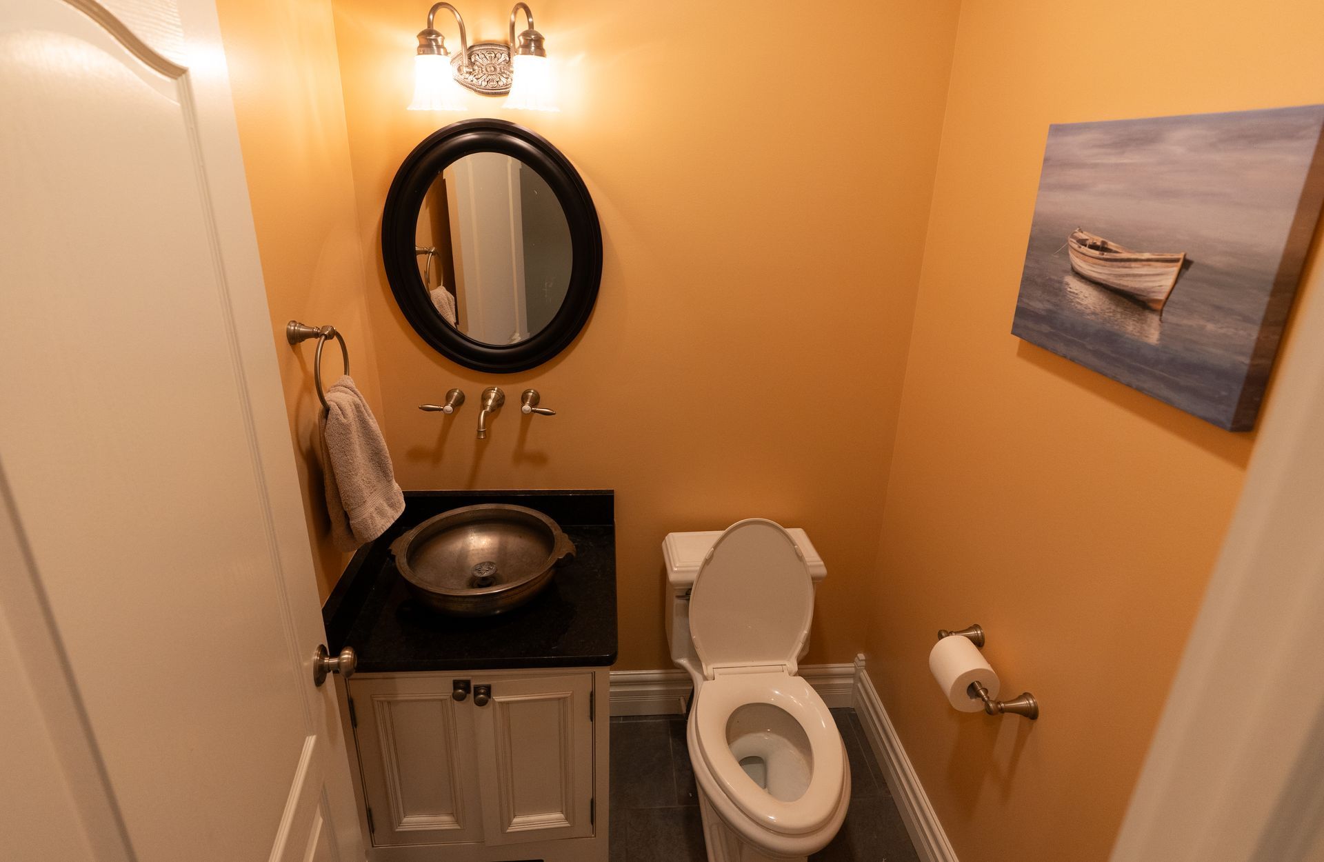 Small bathroom with a toilet, sink, and mirror. The walls are orange, and there is a boat painting.