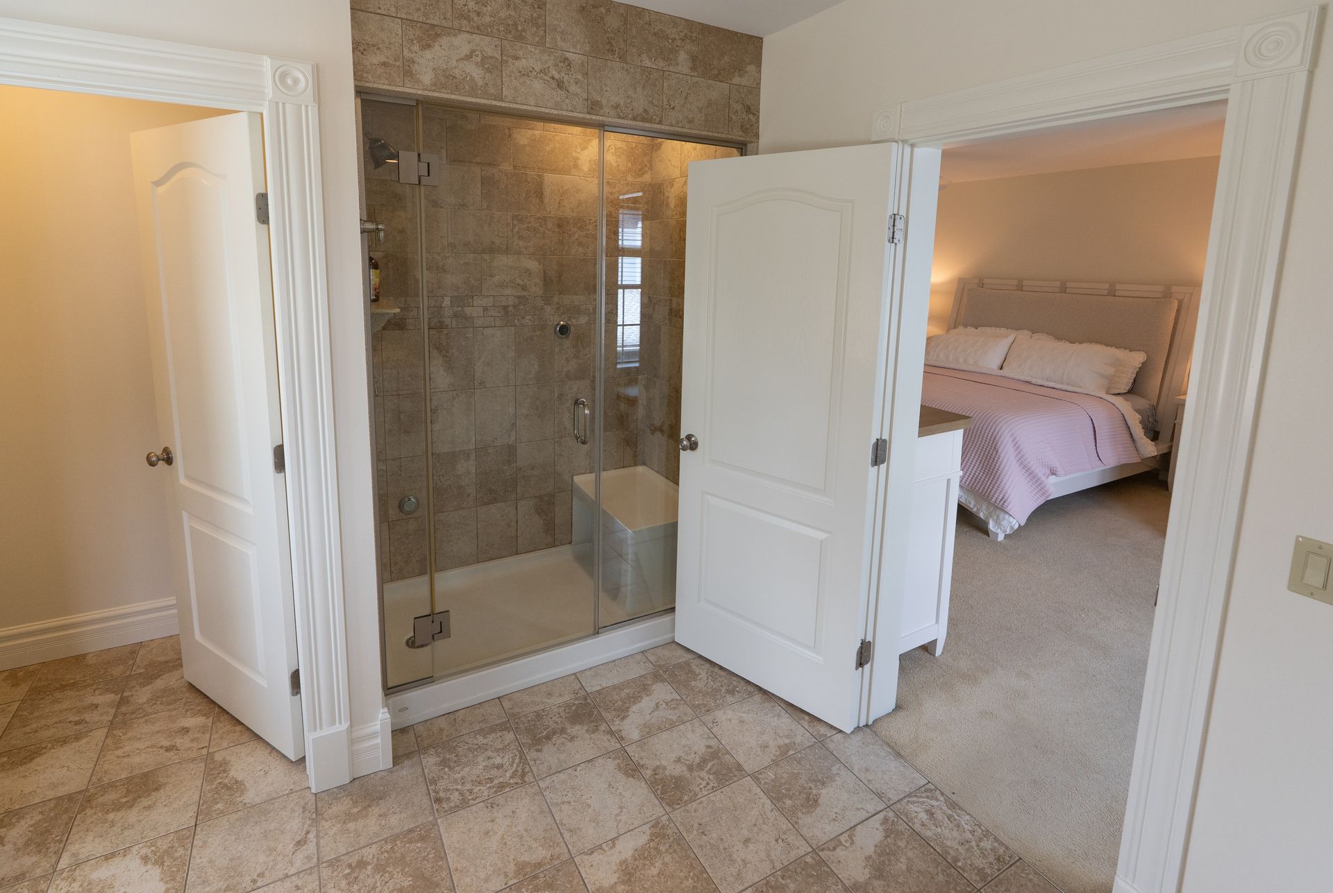 Bathroom with a shower, two doors, and a view of a bedroom. Tile floors and walls, white doors.