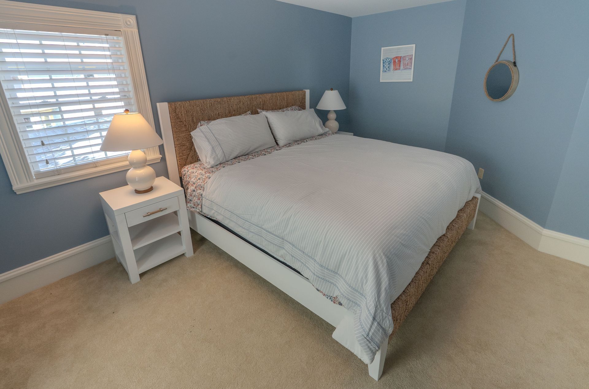 Bedroom with a double bed, nightstand, lamp, and window with blinds. Blue walls, beige carpet.