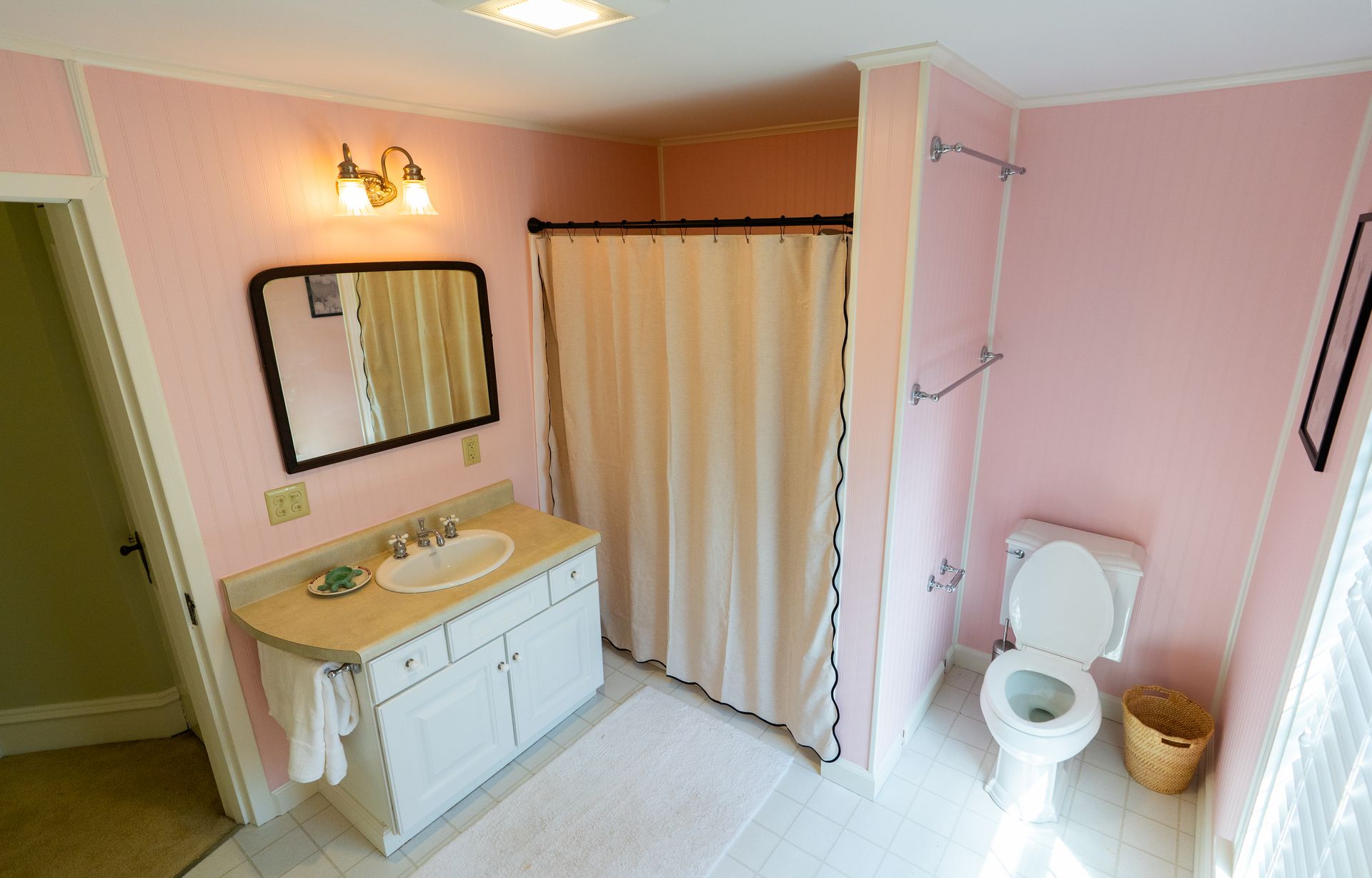 Pink-walled bathroom with a white vanity, toilet, and shower curtain; a gold-framed mirror hangs above the sink.