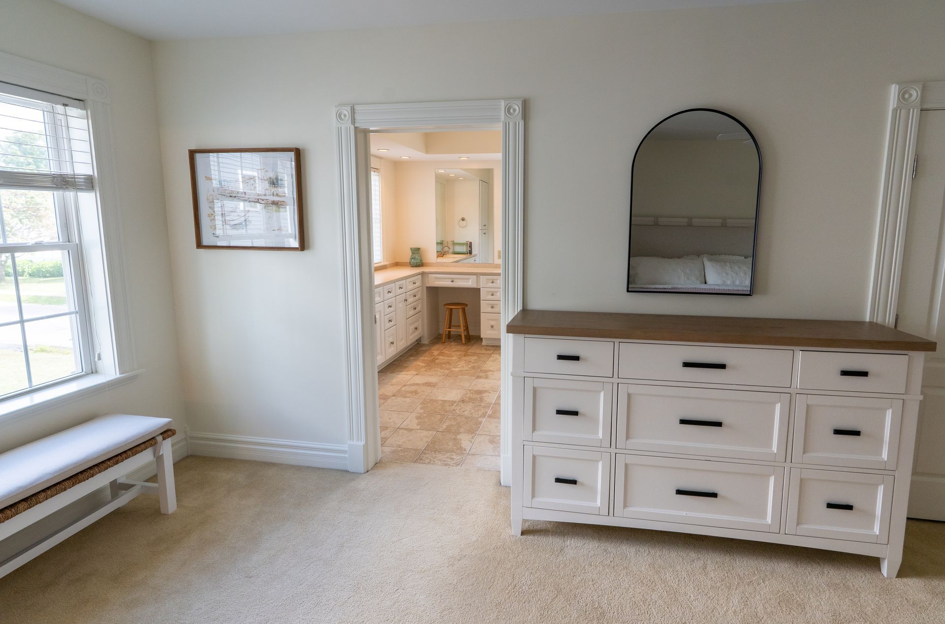White bedroom with dresser, arched mirror, bench, and doorway to a bathroom.