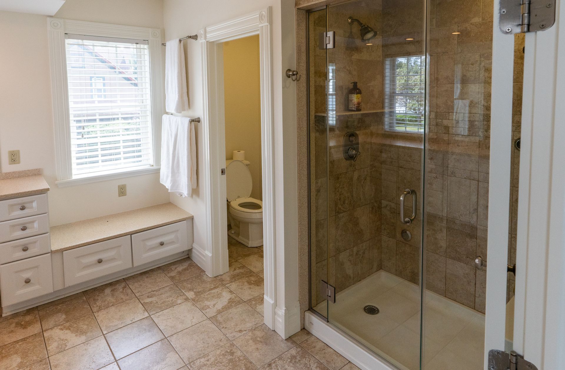 Bathroom with a glass-door shower, toilet, built-in bench, window, and towel rack. Brown tile floors, light walls.