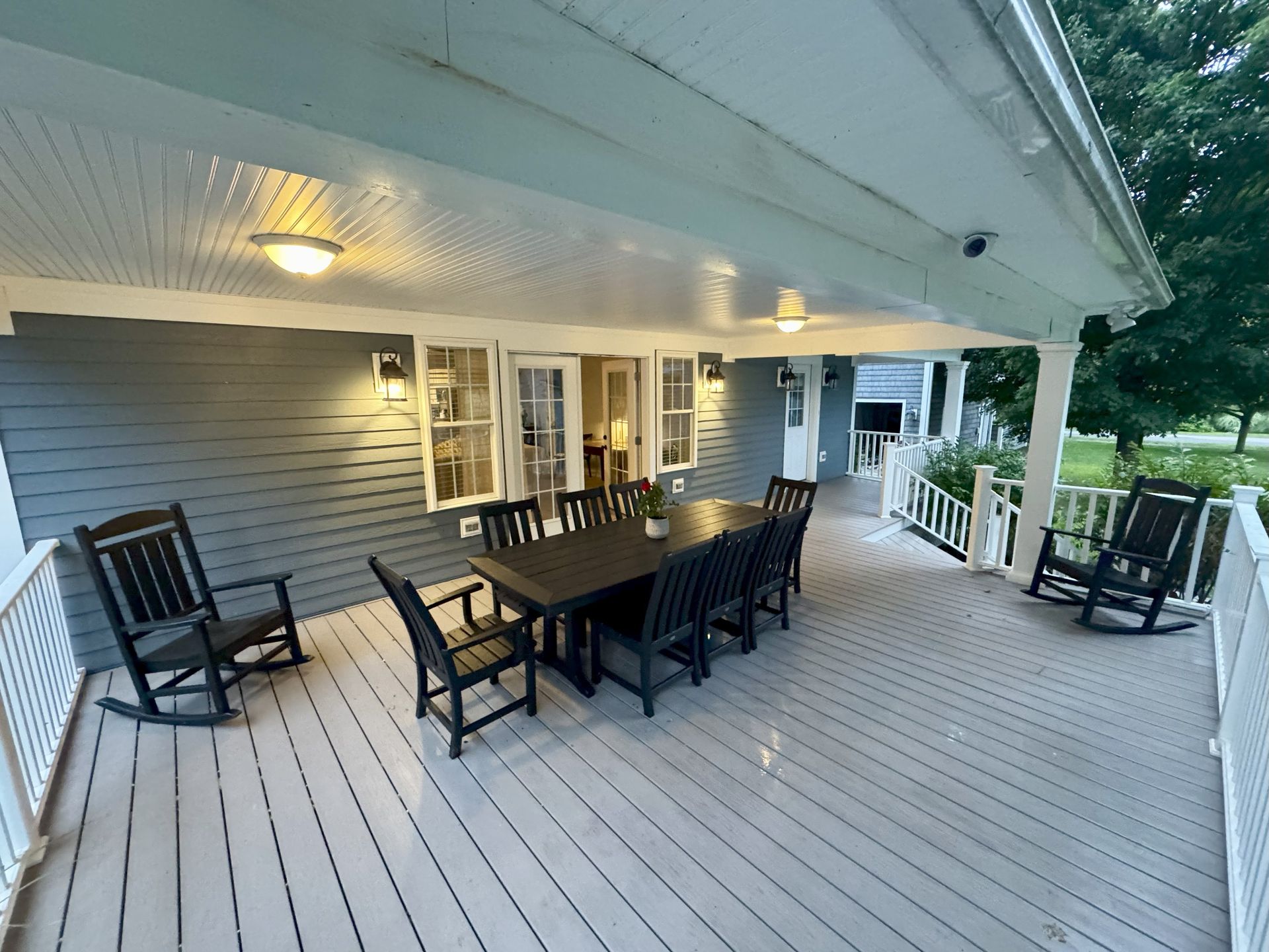 Covered deck with gray siding, a large dark table and chairs, and rocking chairs.