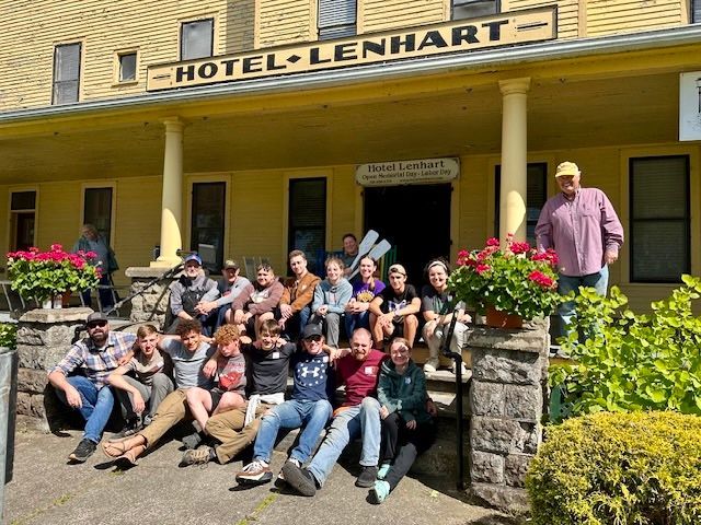 Group of people posing on steps outside Hotel Lenhart. Yellow building with flowers and porch.