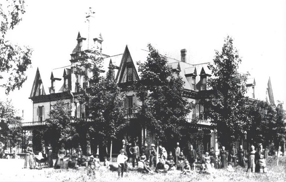 Large Victorian building with group of people in front. Trees partially obscure the building.