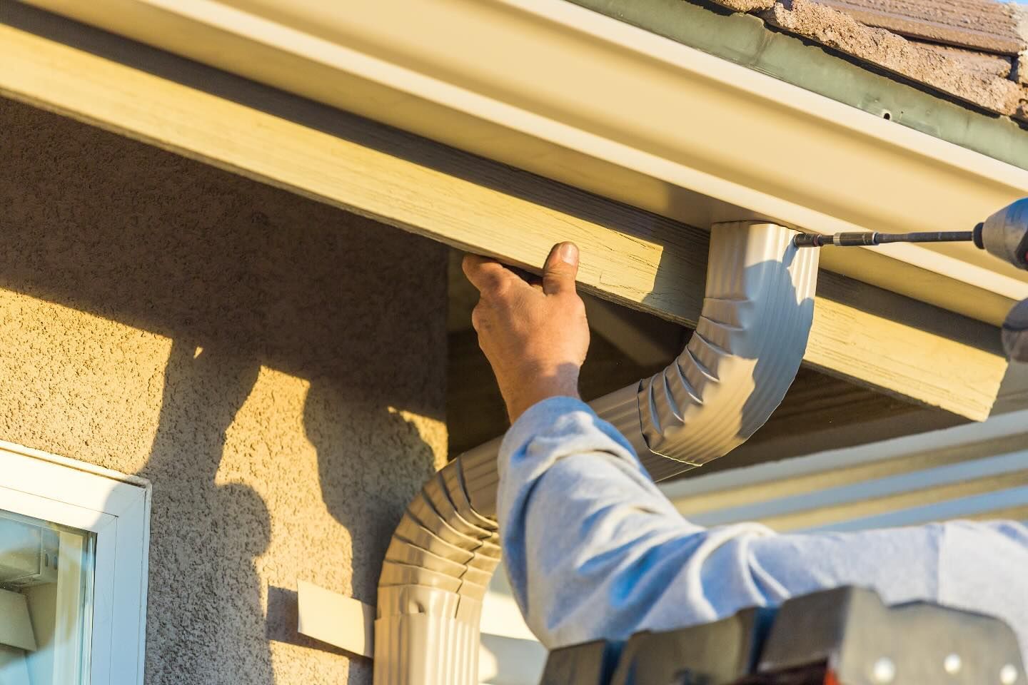 Person inspecting gutter and downspout on a house exterior with a tool
