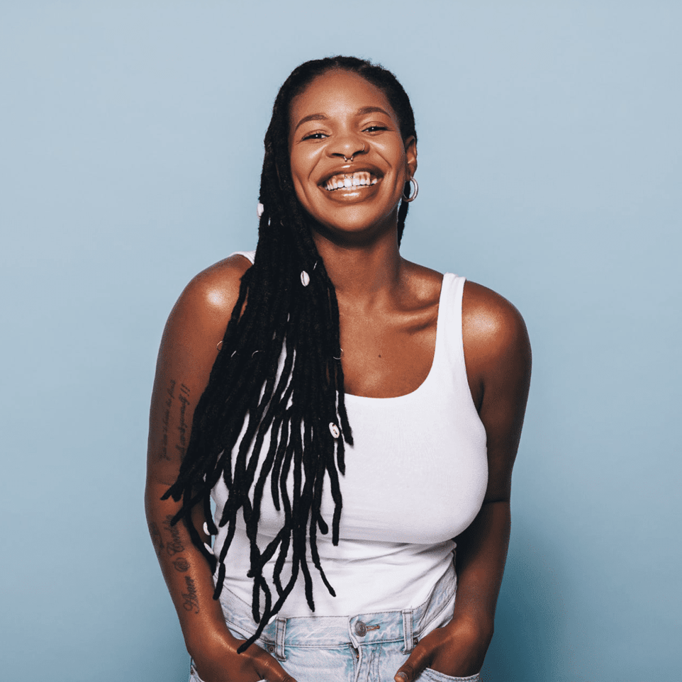 A woman with dreadlocks is wearing a white tank top and smiling.