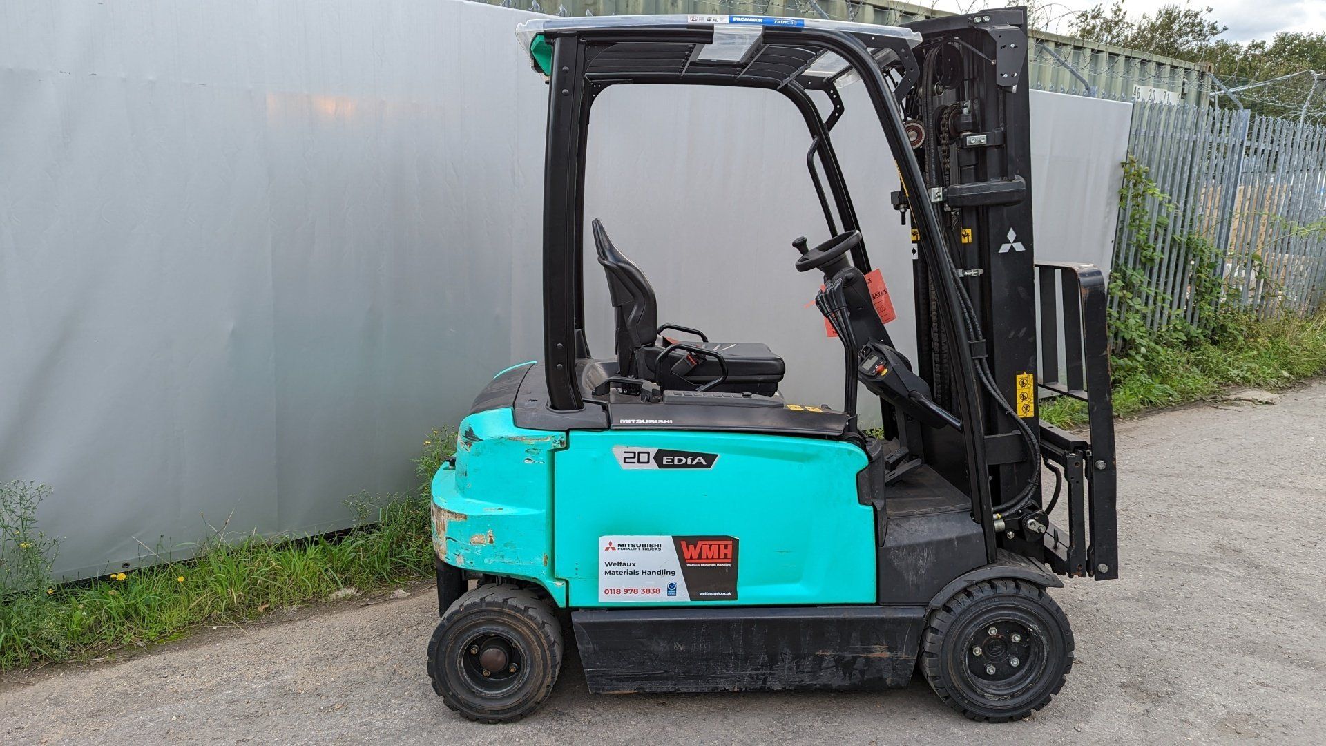 A blue forklift is parked on the side of the road next to a fence. Mitsubishi