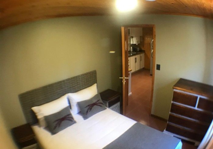 Un dormitorio con una cama, mesita de noche, tocador y puerta que da a una cocina.