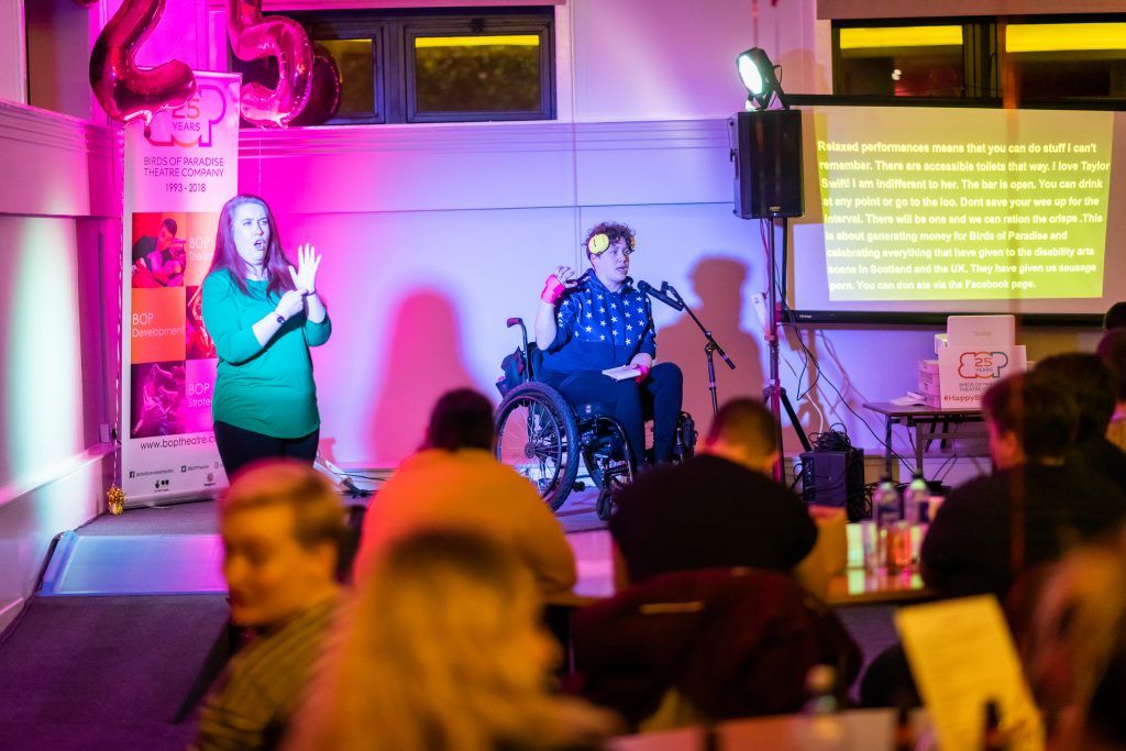Performer on wheelchair on stage with BSL interpreter and captioned screen.