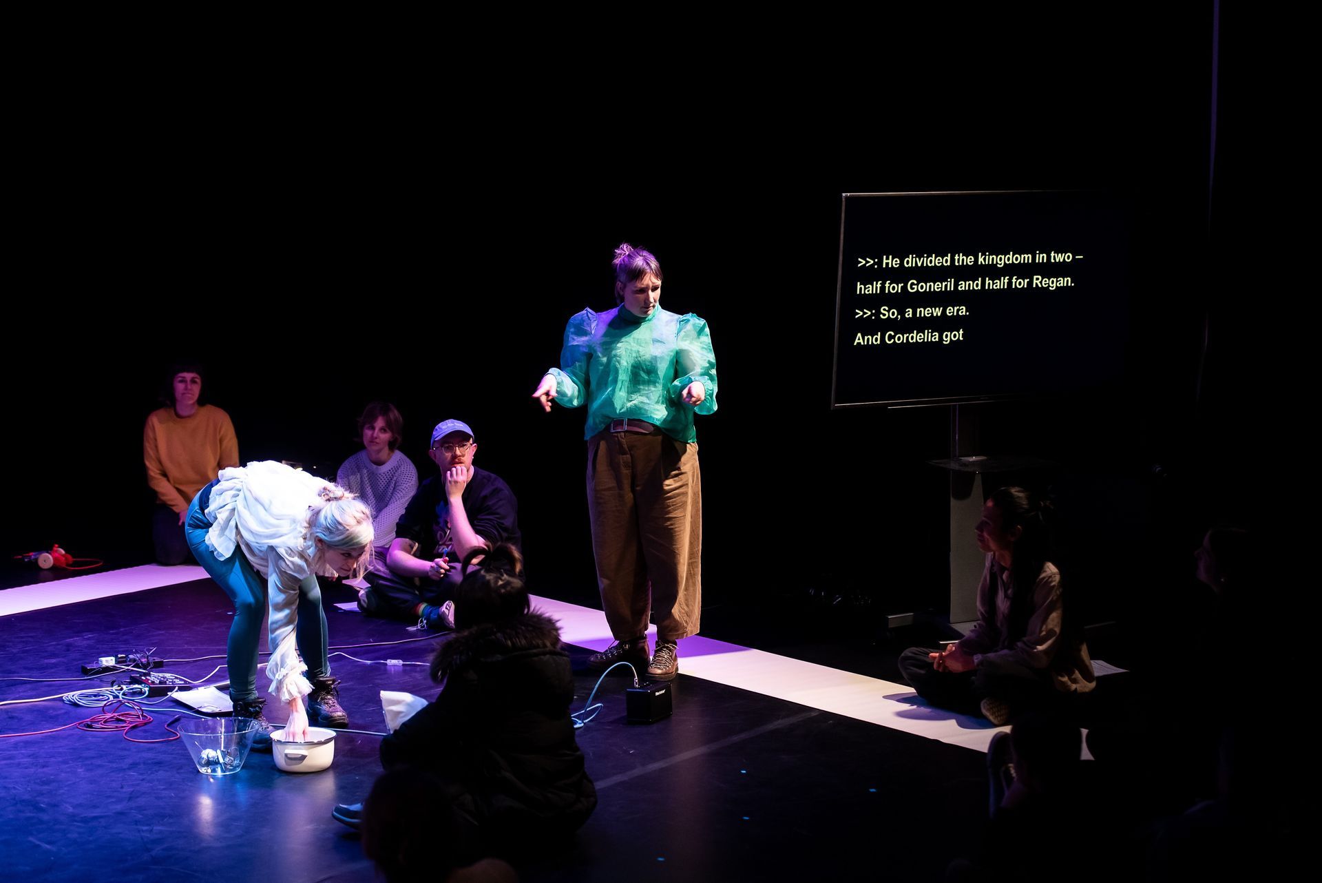 Performer on stage, points to something in foreground with both hands and a quizzical look.  They are surrounded  by seated audience members on the stage.  To one side, is a television screen with yellow captions.