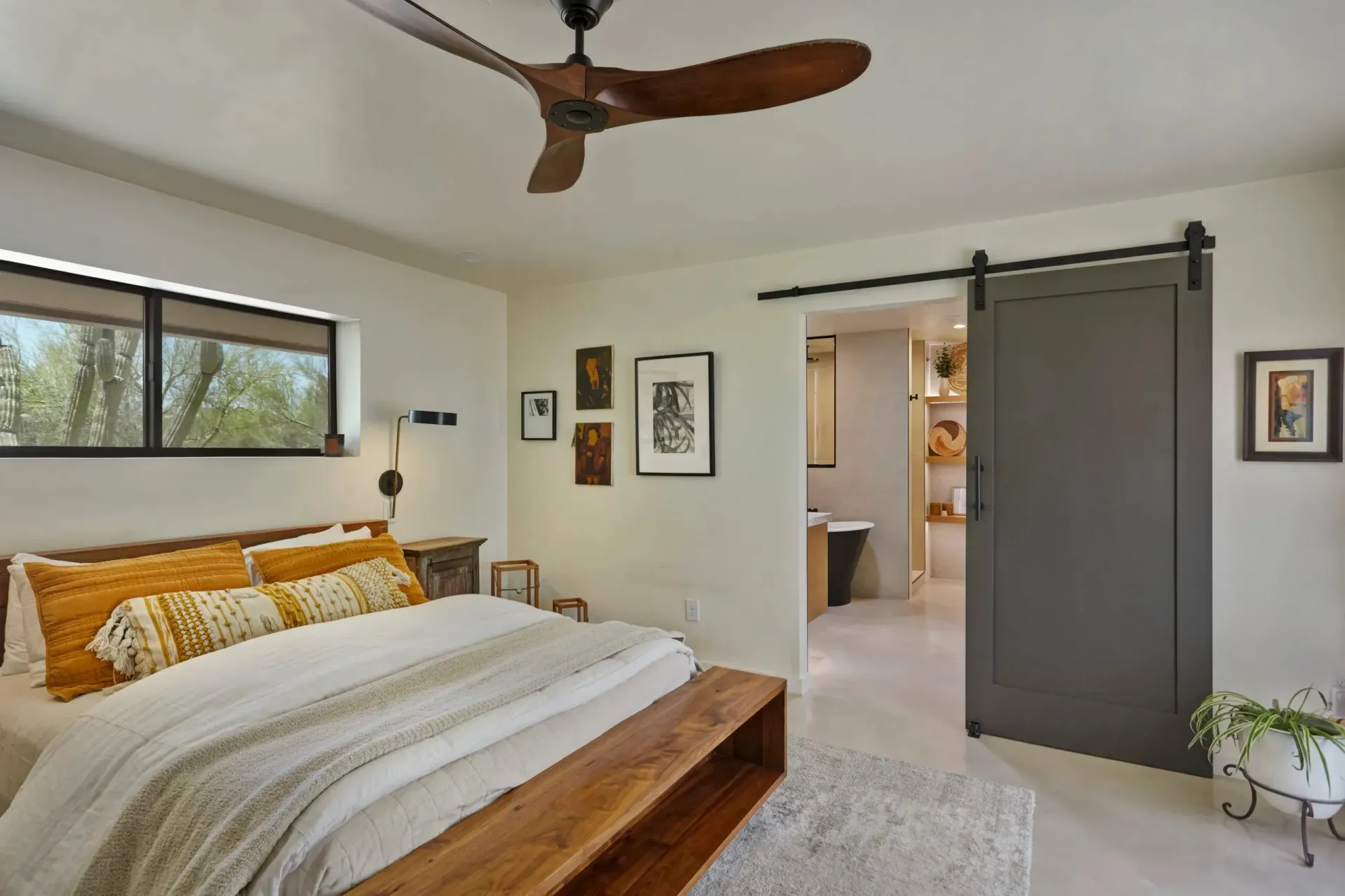 Bedroom with bed, bench, and sliding door to bathroom. Includes ceiling fan, art, and plants.