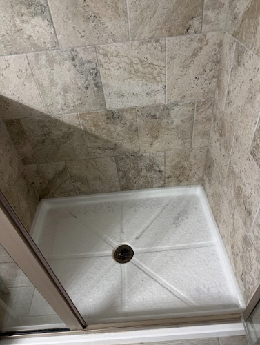 White shower pan with a central drain, surrounded by beige tiled walls. A partially open glass door is visible.