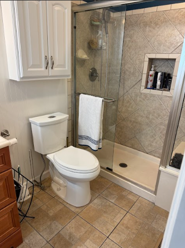 Small bathroom with a toilet, shower, and cabinet. Beige tile floor and shower walls. White toilet, cabinet, and shower base.