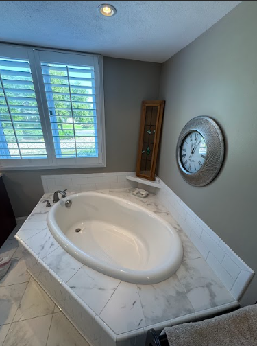 An oval tub in a marble surround, with a clock, small cabinet, and a shuttered window.
