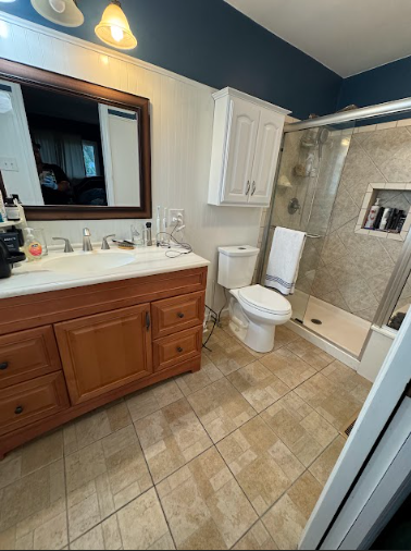 Bathroom with wood vanity, white toilet, and tiled shower with a glass door. Blue and white walls, beige floor tiles.