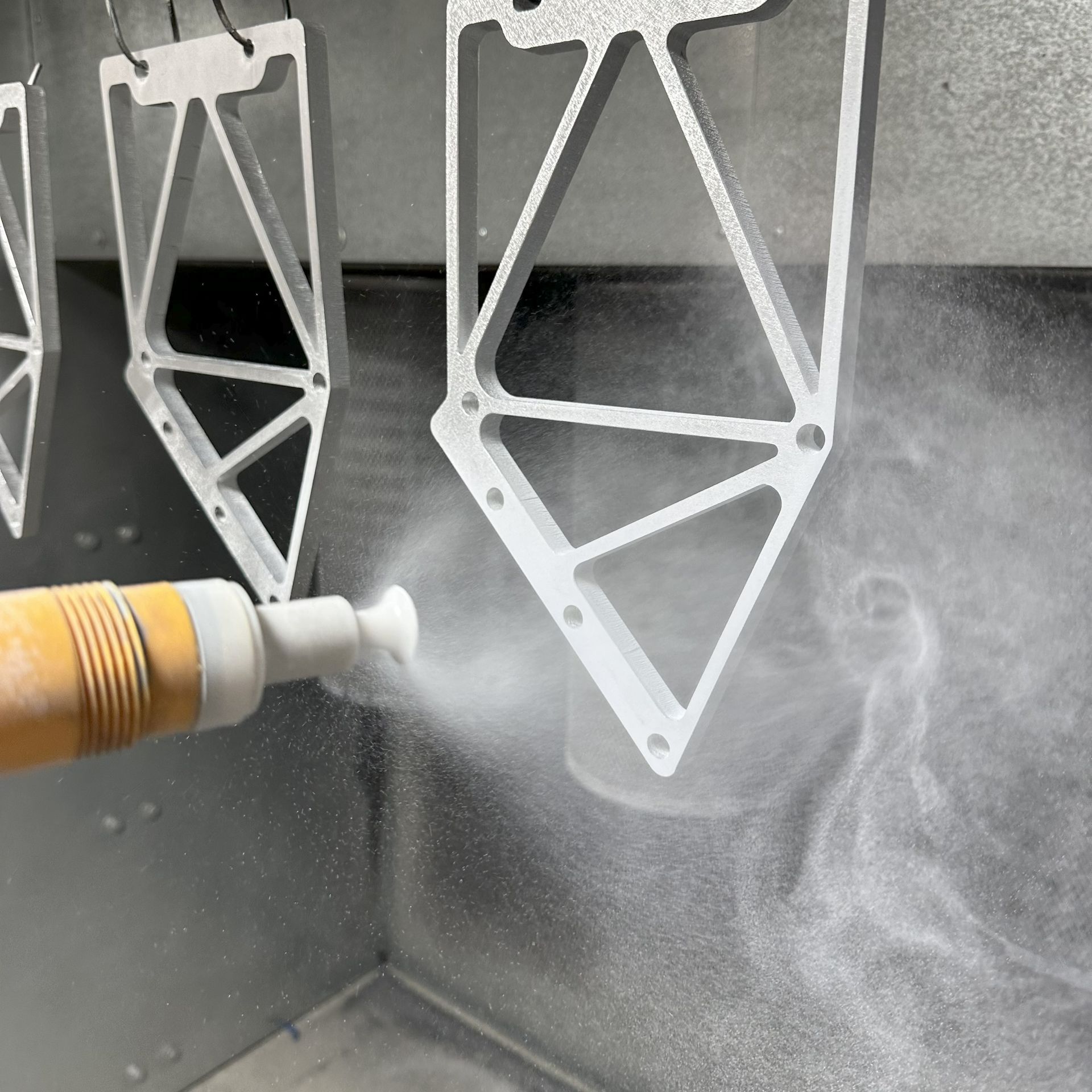 Powder coating aluminum parts with a white spray gun inside an industrial booth.