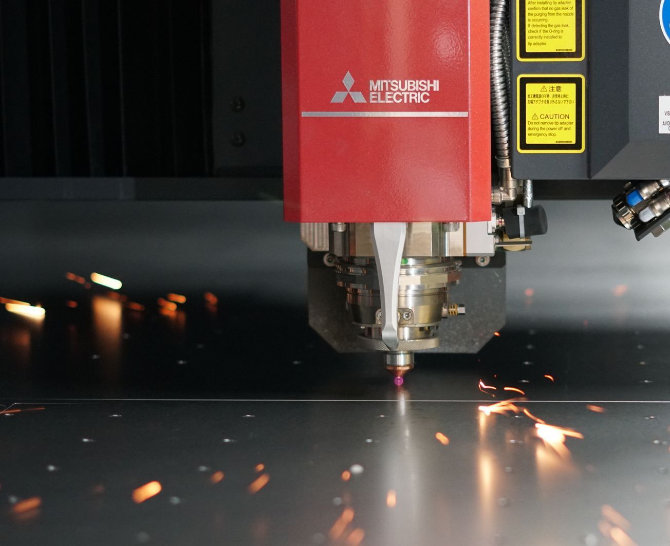 A laser cutting machine by Mitsubishi Electric cuts through metal, creating sparks. Red housing in a factory.