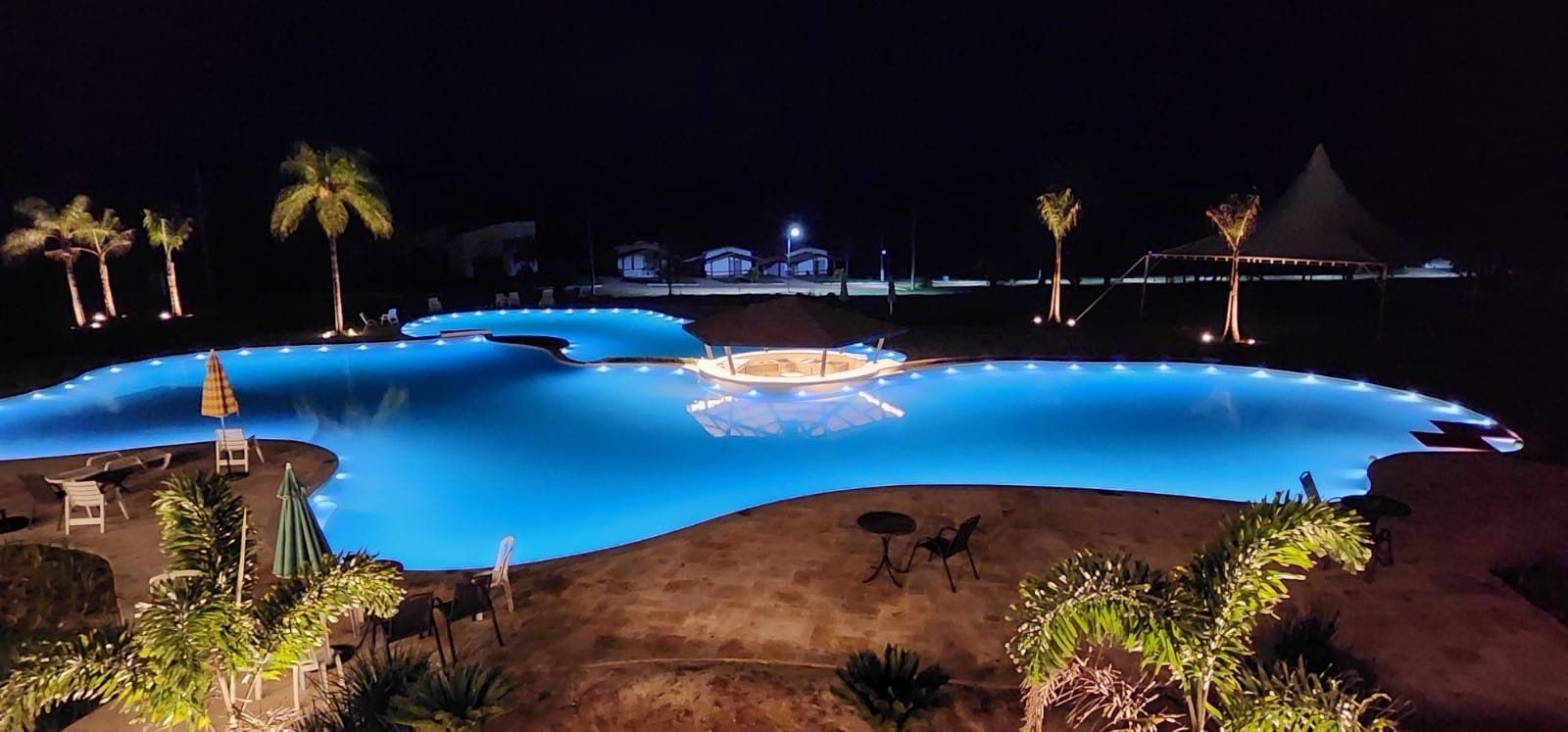 Uma grande piscina é iluminada à noite com palmeiras ao fundo
