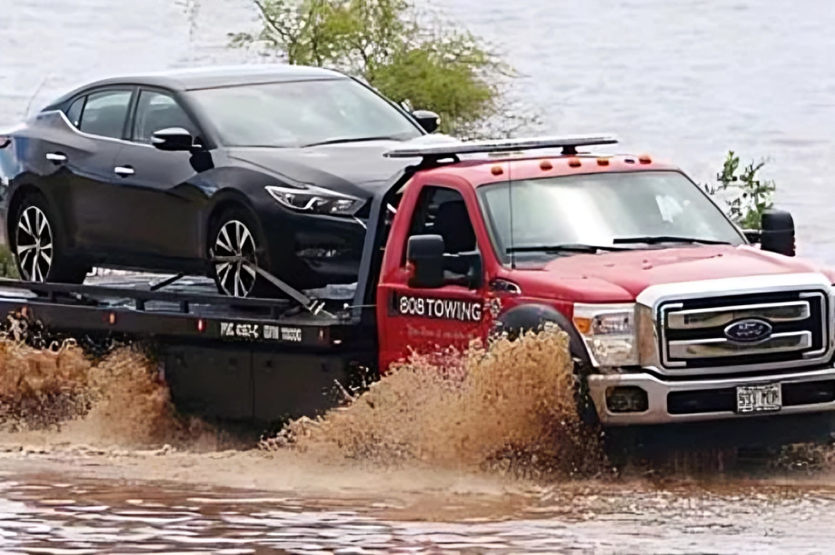A red tow truck is towing a black car through a flooded area.