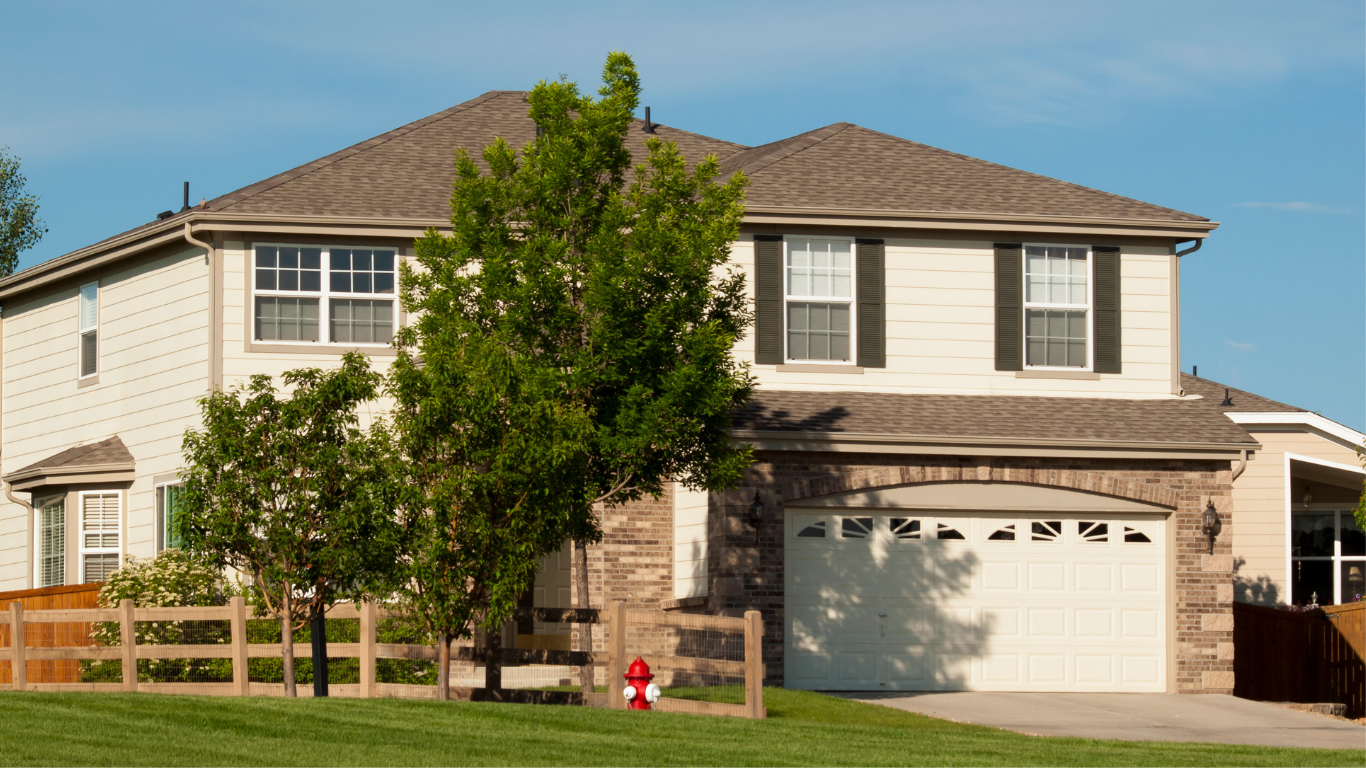 Two-story beige house with brown roof, attached garage, and a small tree in front.