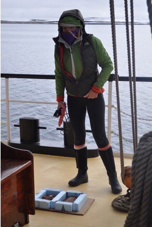 A woman wearing a green jacket and black pants is standing on a boat