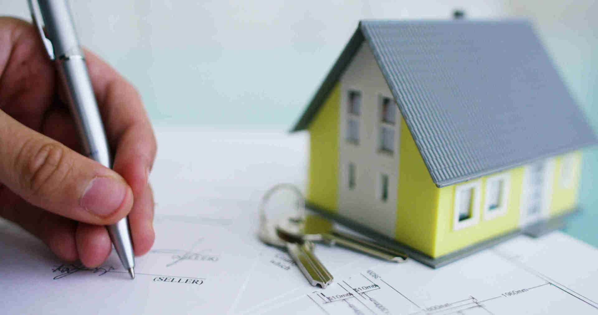 Eine Person schreibt auf ein Stück Papier neben einem Modellhaus und Schlüsseln.