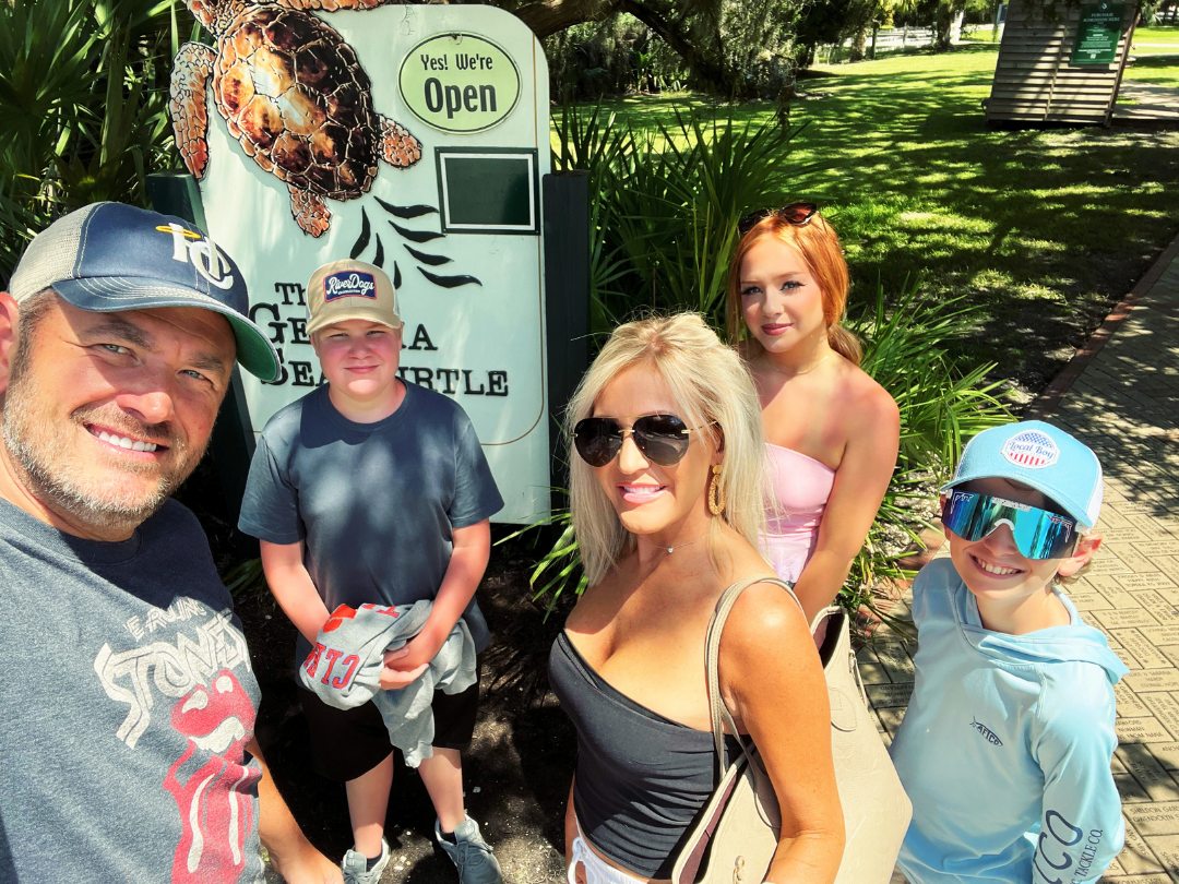 Family of five poses in front of a sign for a turtle sanctuary outdoors on a sunny day. Family of five poses in front of a sign for a turtle sanctuary outdoors on a sunny day.