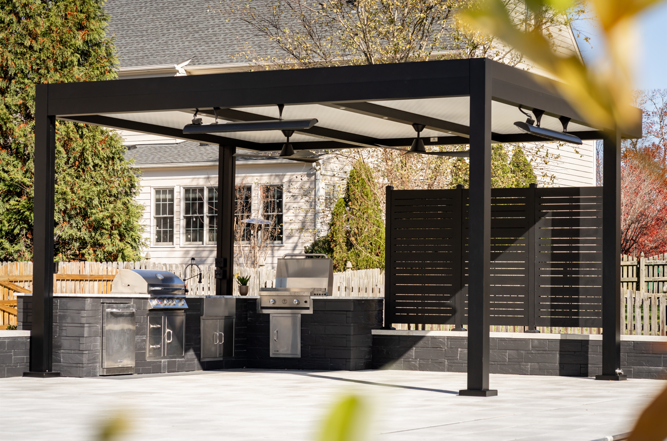 Outdoor kitchen with black pergola, grill, fans, and built-in counter, backyard setting.