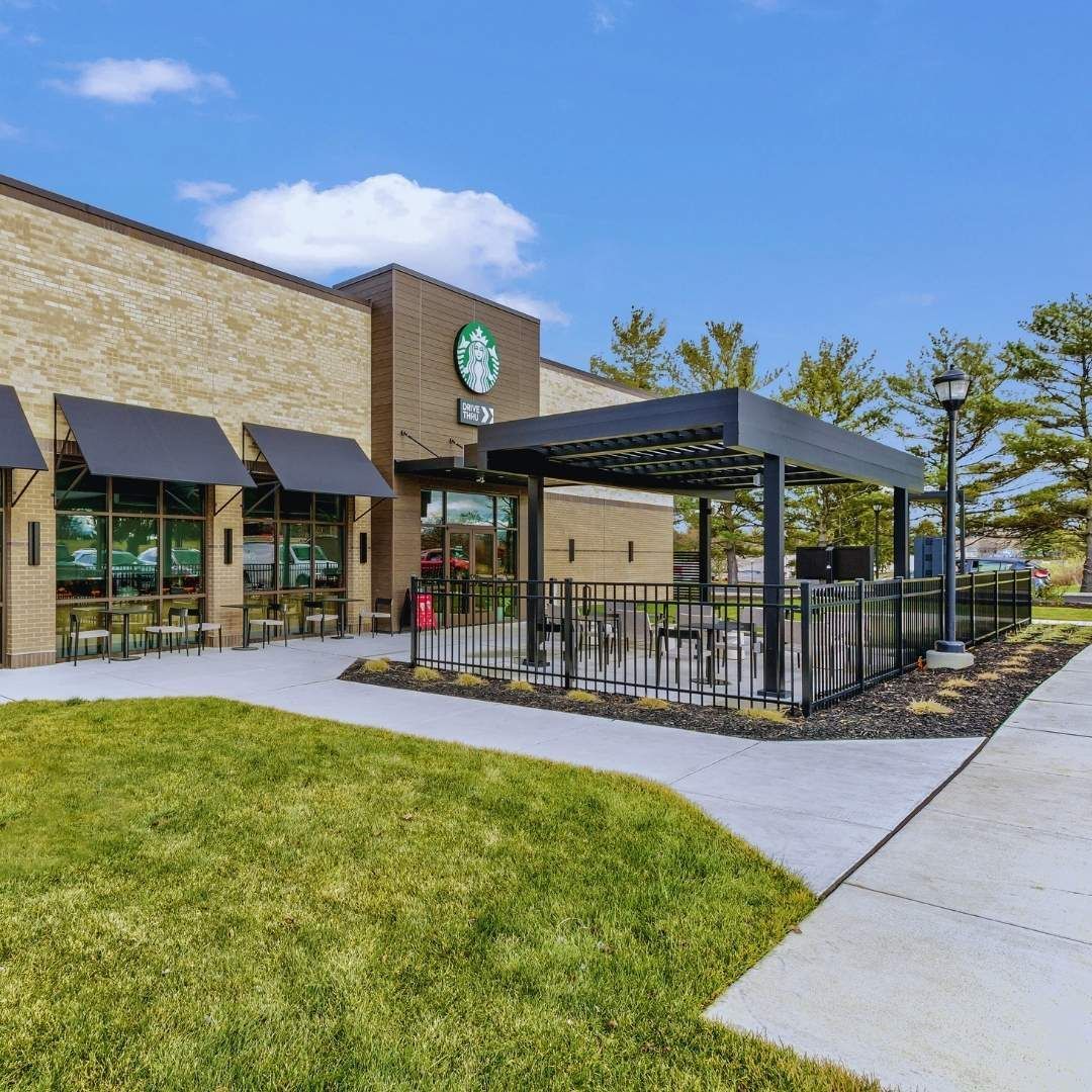 Starbucks store with outdoor seating, green lawn, beige brick exterior, and blue sky.