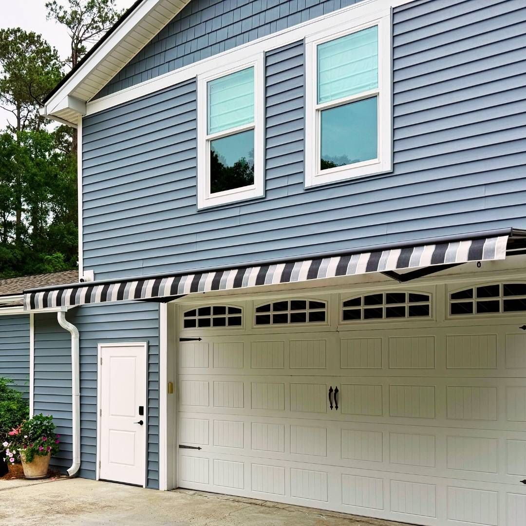 Blue-sided house with white garage door; black and white striped awning; white door; trees in background.