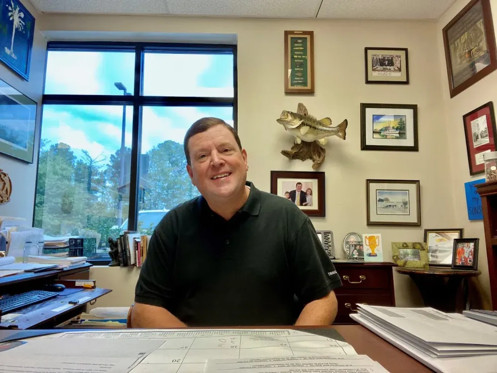 Smiling man seated at an office desk with framed photos and certificates on the wall