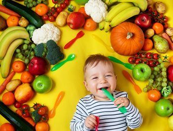 Baby surrounded by colorful fruits and vegetables, smiling while holding a green spoon on a yellow background.