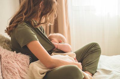 Woman seated, breastfeeding baby indoors. Sunlight illuminates.