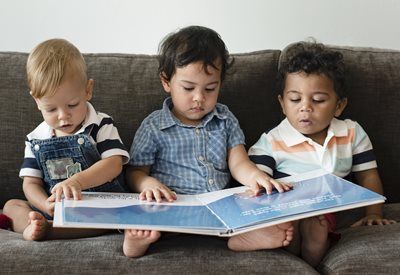 Three young children sitting together reading a book on a gray couch.