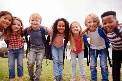 Group of smiling children with backpacks, arms around each other, outdoors.