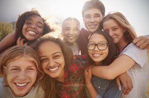 Group of smiling teens, arms around each other, outside on a sunny day.
