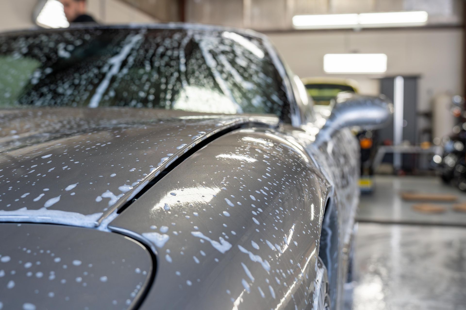 Silver car covered in soapy foam, being washed inside a garage.