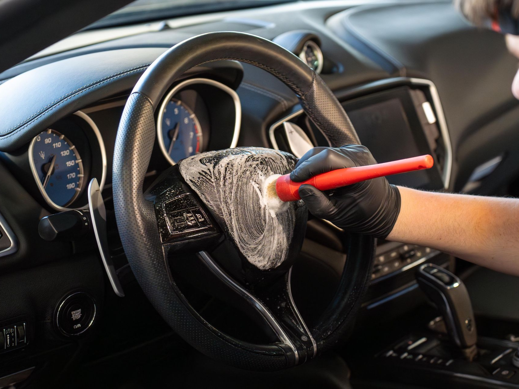 Person cleaning a car's steering wheel with a brush and cleaning solution. Black gloves, dark interior.
