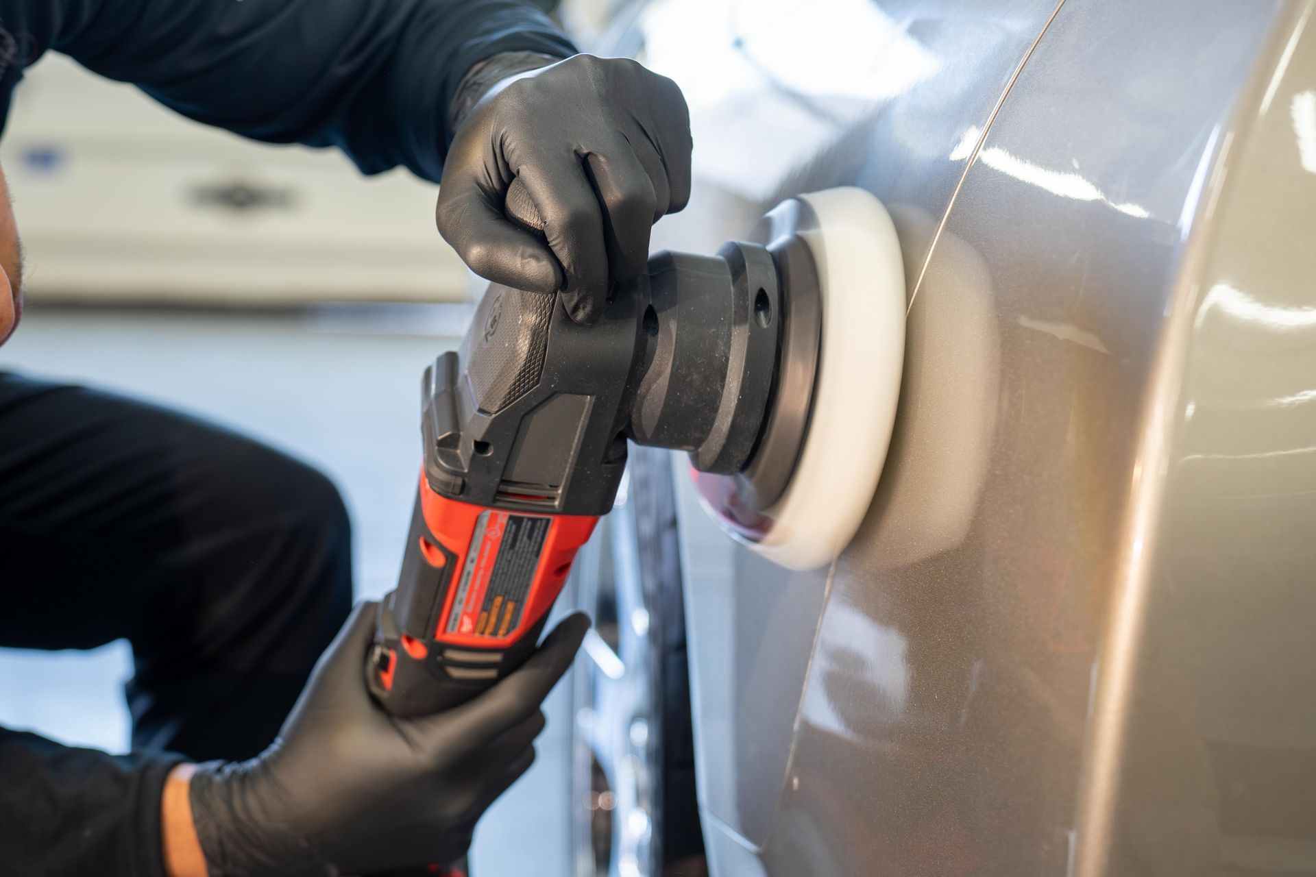 Person in black gloves using a power polisher on a car's side panel, buffing the light-colored surface.