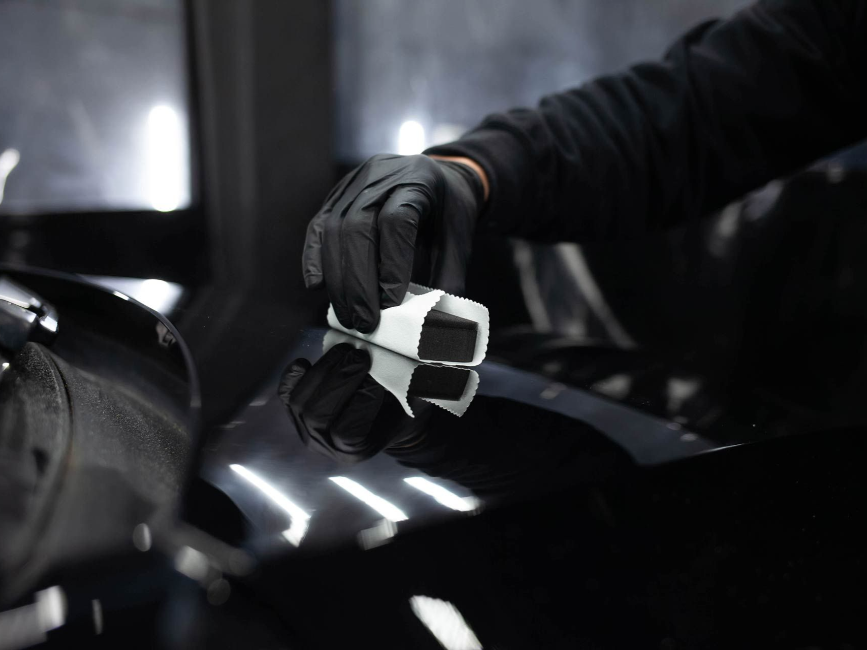 Gloved hand applying a ceramic coating to a glossy black car hood in a well-lit shop.