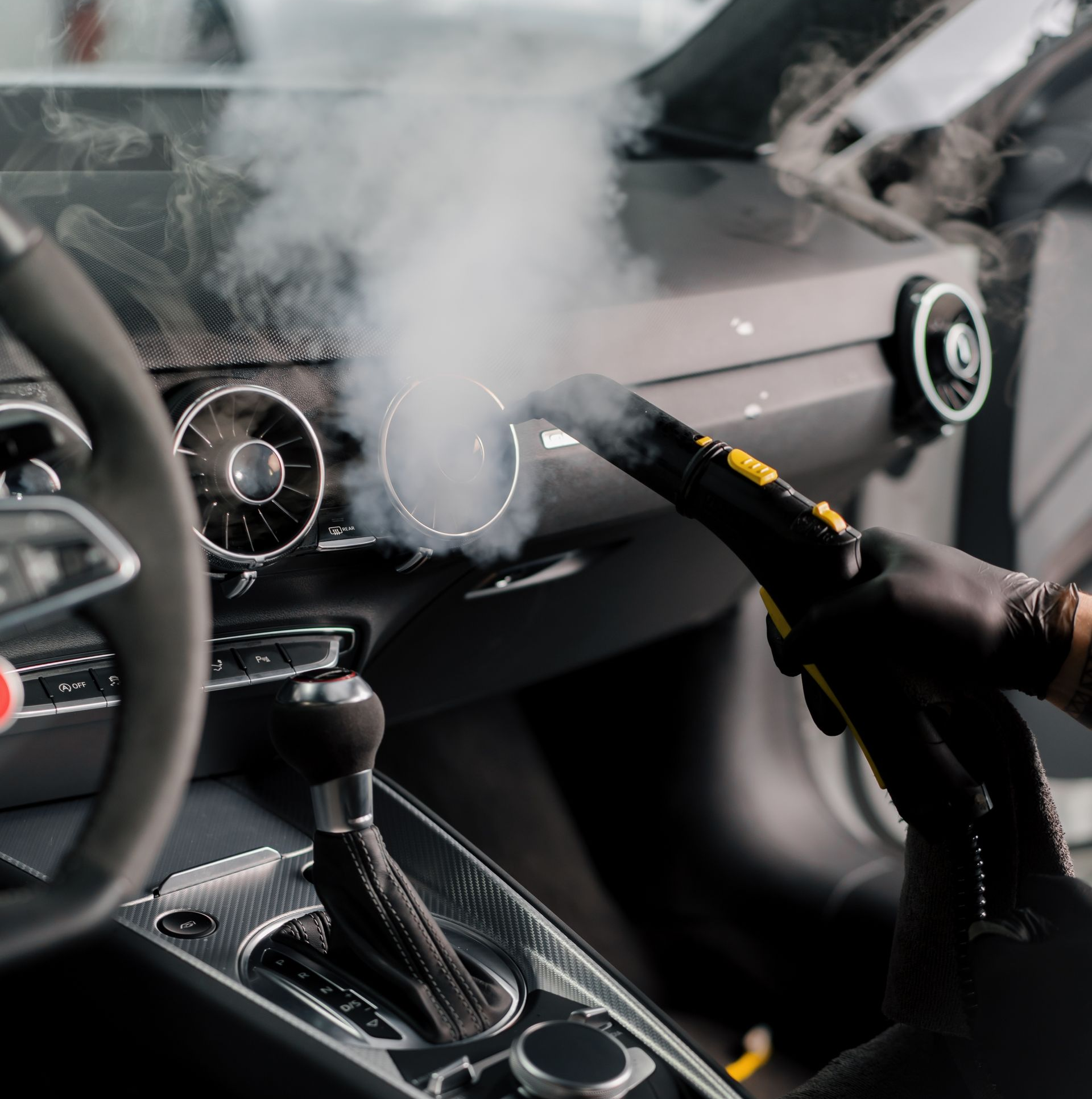 Car interior being steam cleaned, focusing on dashboard vents with steam visible.
