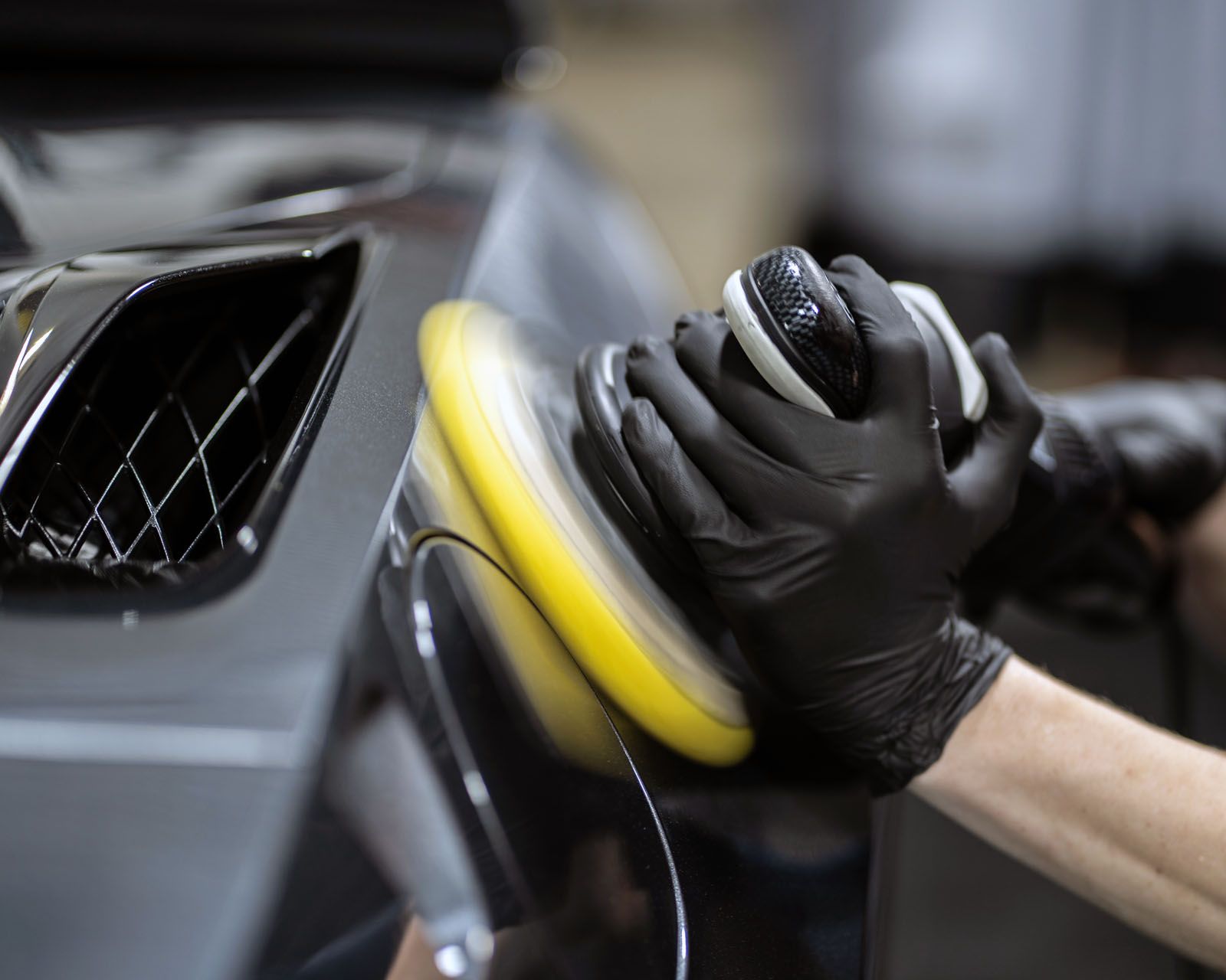 Person using a buffing machine on a dark gray car, wearing black gloves.