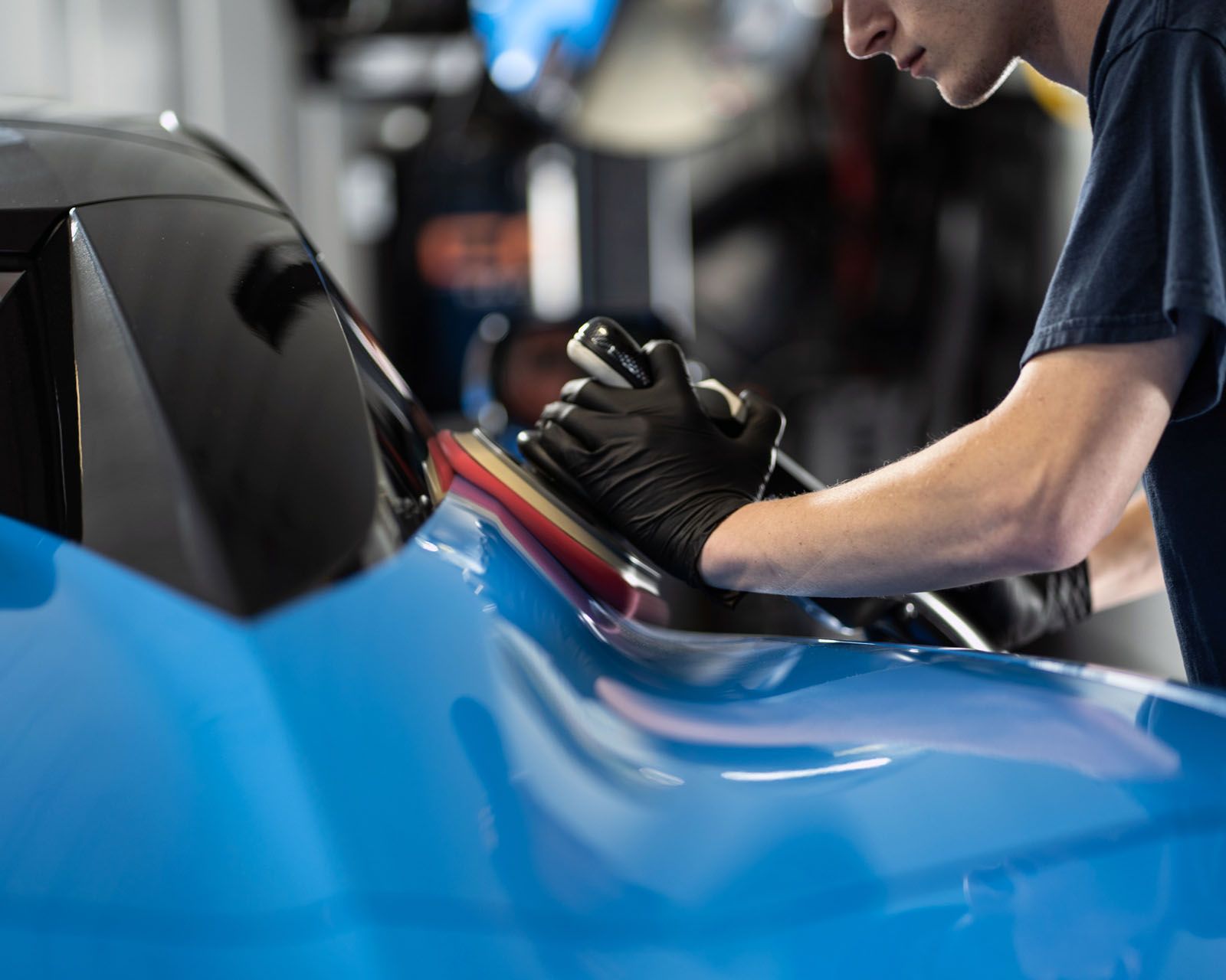 Person in black gloves polishing a blue car, using a handheld polisher.